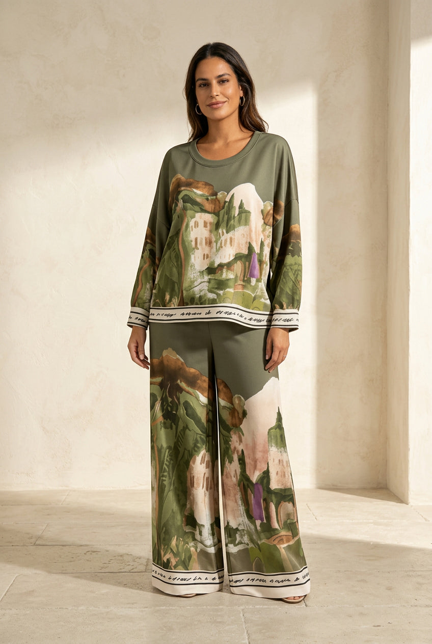 A woman modeling a matching two-piece lounge set with a landscape print. The set includes a long-sleeve top and wide-leg pants in olive green with a scenic print and white striped cuffs and hem.
