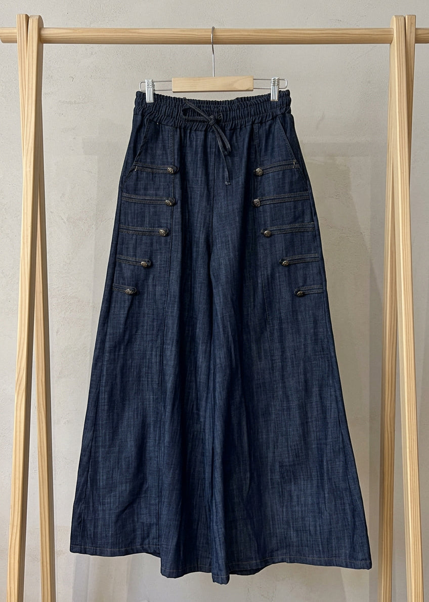 A pair of dark blue denim wide-leg pants hanging on a white hanger against a light beige wall. The pants feature an elastic drawstring waist, wide legs, and decorative buttoned flap pockets on the sides.
