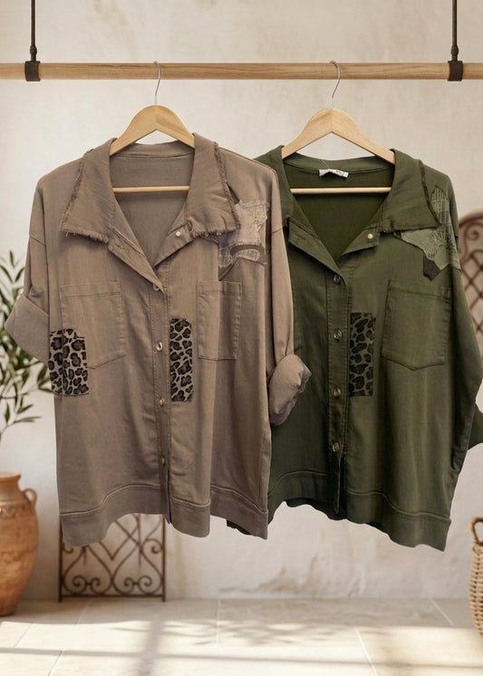 Two patchwork button-up shirts hanging on black hangers against a plain white wall. The left shirt is taupe with leopard-print and camouflage patches, and the right shirt is olive green with similar patches.