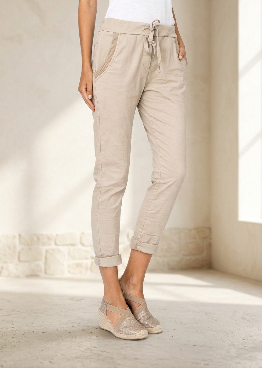 A person wearing light beige drawstring cropped pants with rolled cuffs and beige wedge espadrilles, standing in a sunlit room with cream walls and stone flooring.