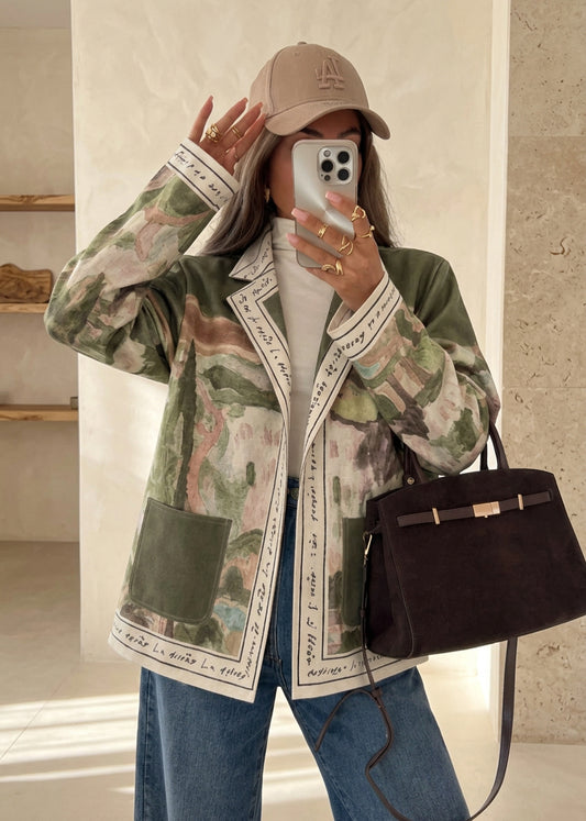 A woman takes a mirror selfie in a modern kitchen setting, wearing a beige baseball cap, a green and beige camouflage print jacket with white script trim, a white turtleneck, blue jeans, and carrying a dark brown leather handbag.