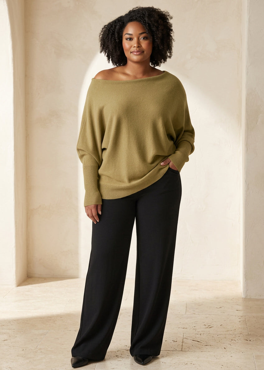 A woman modeling a loose, off-the-shoulder olive green sweater paired with black wide-leg pants, standing against a light gray studio background.
