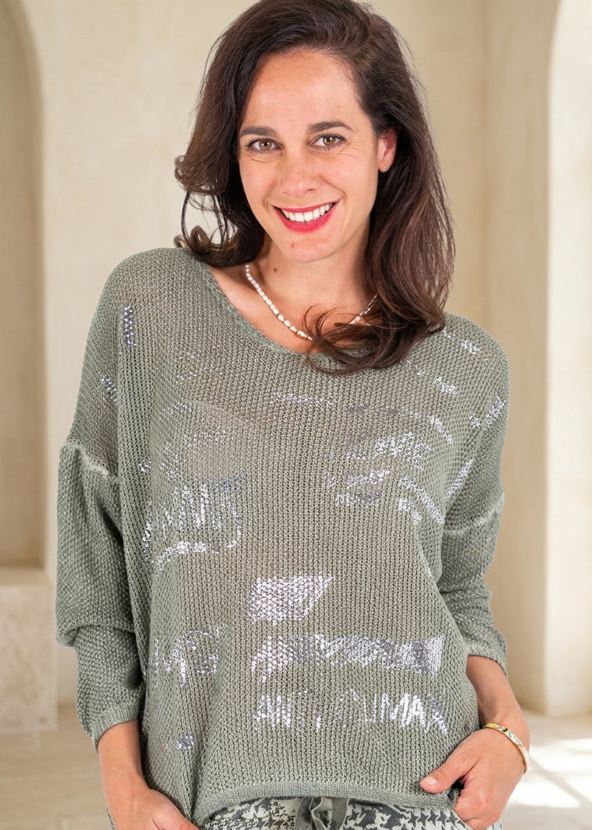 A woman wearing a loose, olive green knit sweater with distressed silver graphic text overlays, paired with a black and white houndstooth patterned top, against a plain white background.