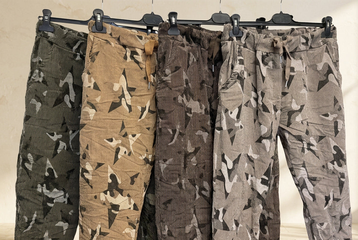 Four pairs of camouflage cargo pants hanging on a black rack against a plain light background. The pants come in four different camo colorways: olive green, tan, dark brown, and light gray-brown.