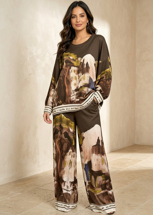 A woman modeling a matching two-piece lounge set featuring a brown abstract landscape print with beige, white, and olive accents, complete with striped cuffs and hems, shot against a light gray studio background.