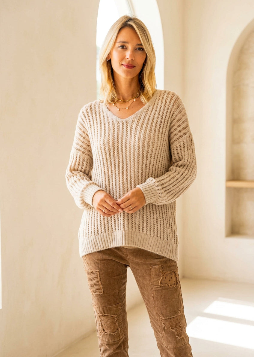 A woman wearing a beige chunky knit V-neck sweater with a textured openwork pattern, paired with brown pants, against a plain white background.