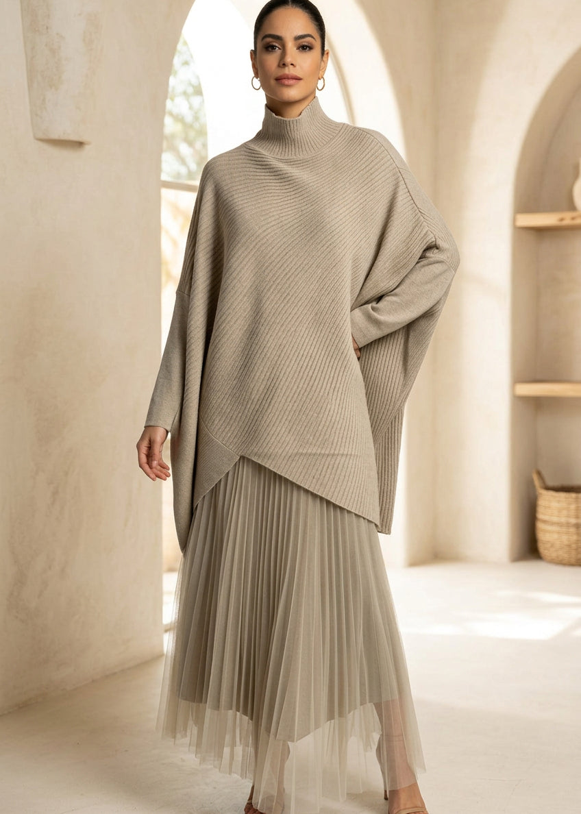 A model wearing a beige ribbed knit turtleneck poncho layered over a matching pleated tulle midi skirt, photographed against a neutral light gray studio background.