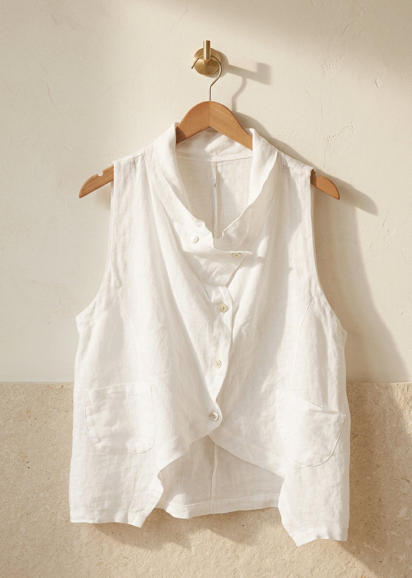 A white sleeveless button-front vest with a relaxed, slightly oversized fit, hanging on a wooden hanger from a gold-toned clothing rack against a solid beige background.
