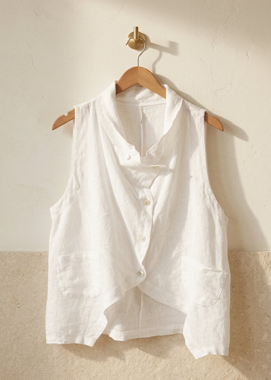 A white sleeveless button-front vest with a relaxed, slightly oversized fit, hanging on a wooden hanger from a gold-toned clothing rack against a solid beige background.
