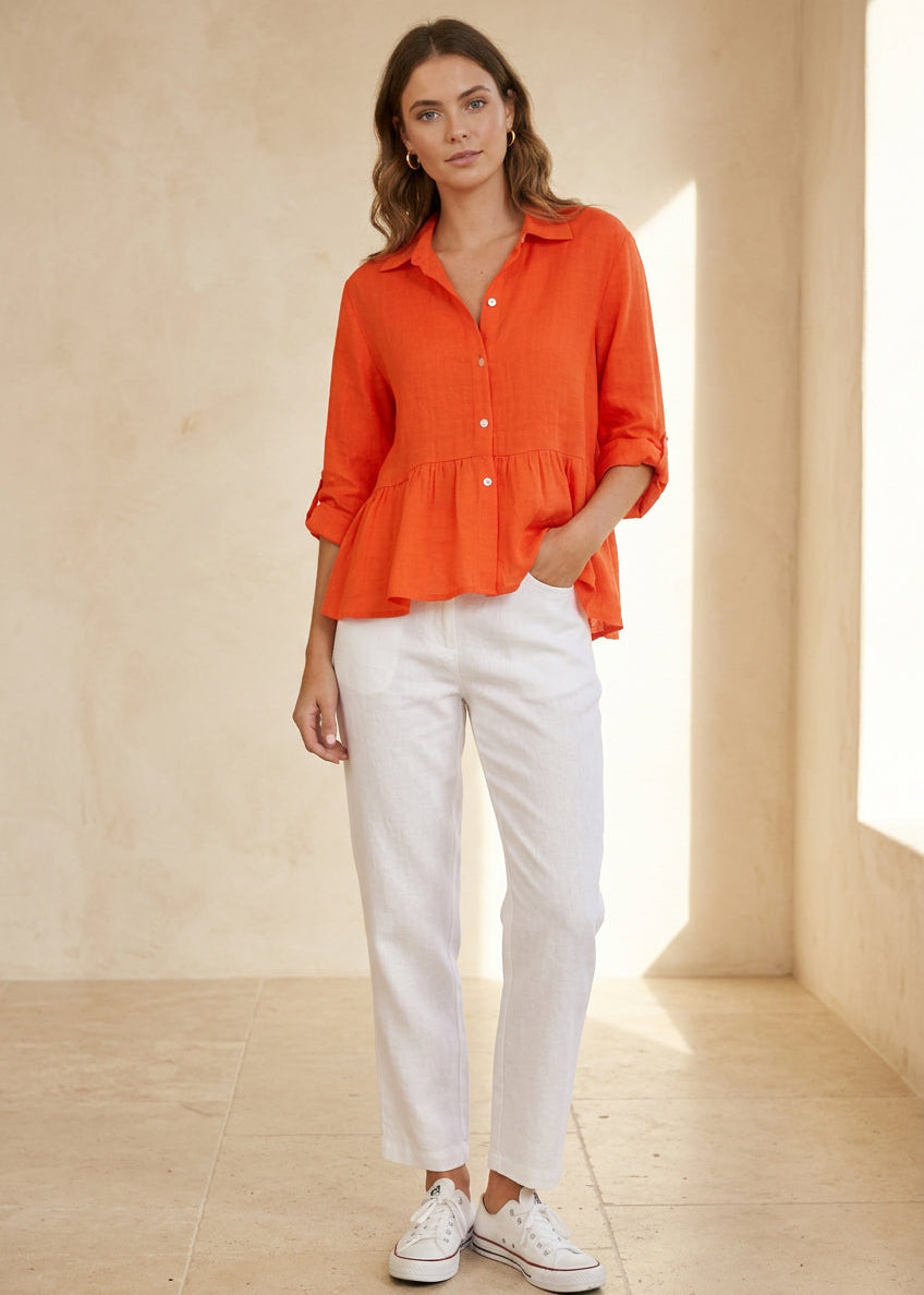 A woman modeling a bright orange long-sleeve button-up shirt with a peplum hem, paired with light-wash blue jeans and white sneakers, standing against a gray tiled wall.