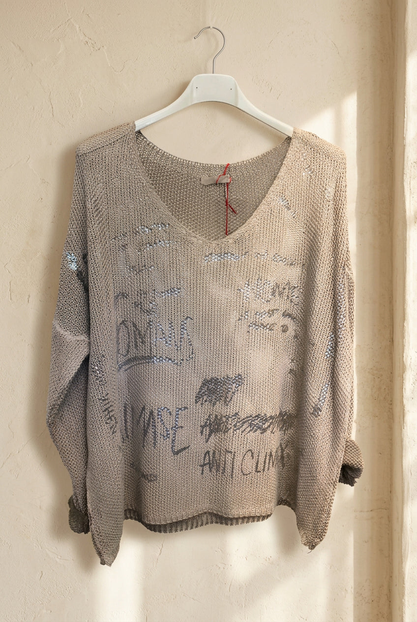A beige chunky knit sweater with sequined graffiti-style lettering, hanging on a white plastic hanger against a white wall.
