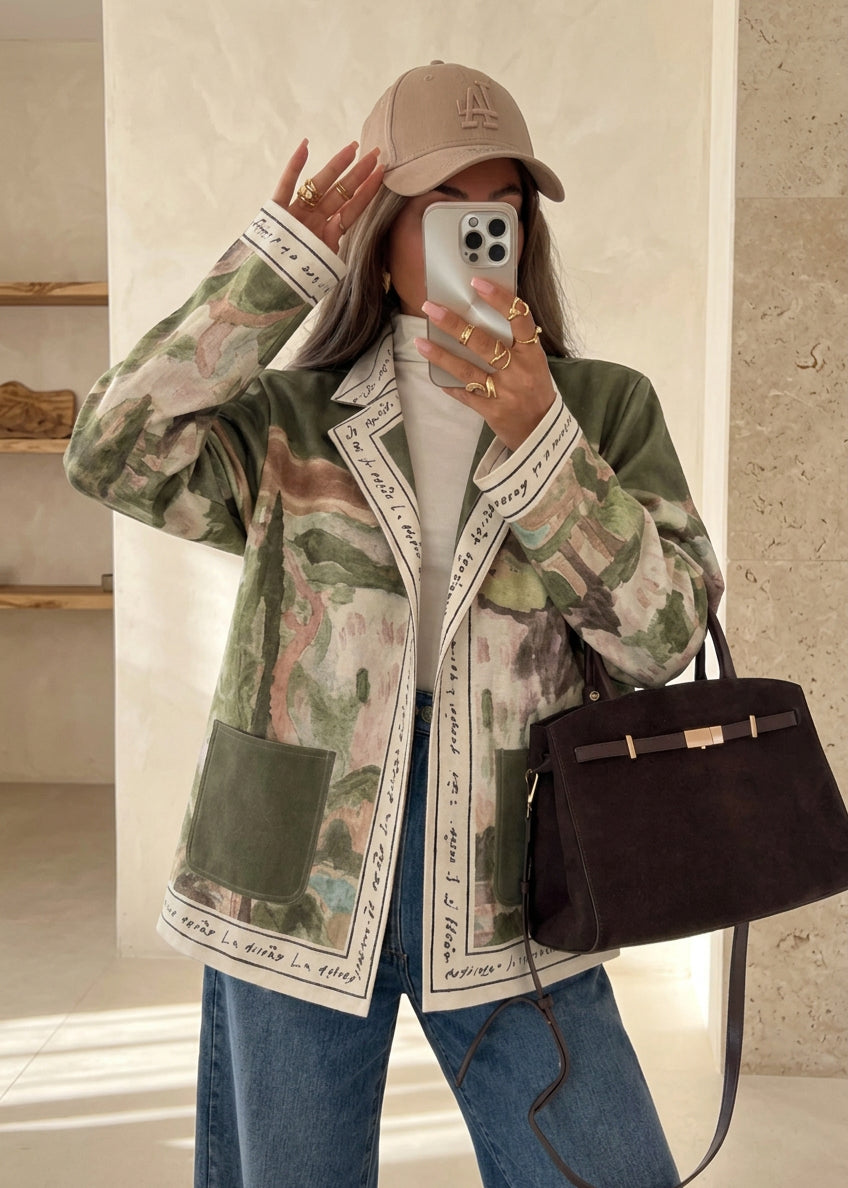 A woman takes a mirror selfie in a modern kitchen setting, wearing a beige baseball cap, a green and beige camouflage print jacket with white script trim, a white turtleneck, blue jeans, and carrying a dark brown leather handbag.