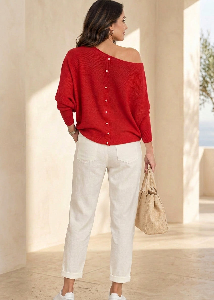 A woman wearing a bright red off-the-shoulder knit sweater with decorative buttons down the center, paired with blue jeans, white sneakers, and holding a black handbag, photographed against a solid gray studio background.