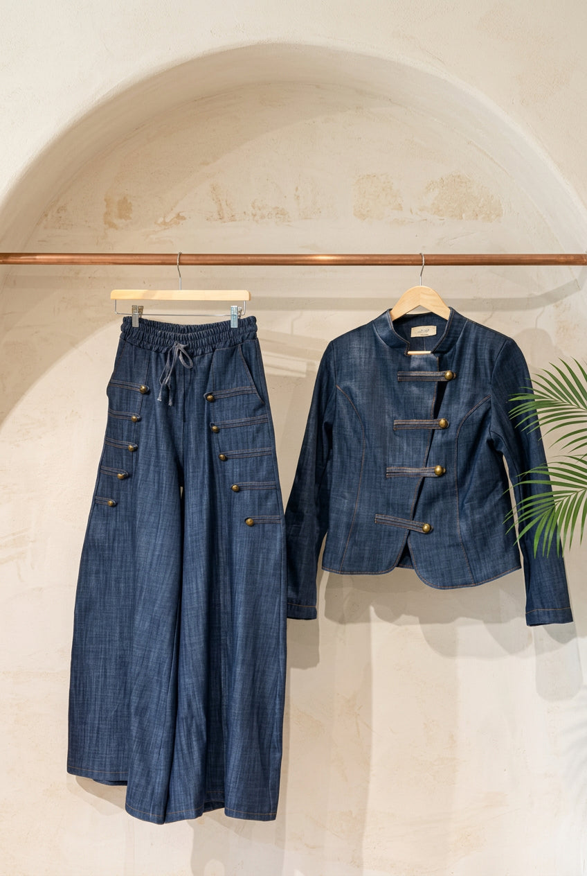 A dark blue denim two-piece outfit hanging on a wooden rack against a warm beige plaster wall. The set includes wide-leg pants with decorative button details on the legs and a matching cropped jacket with toggle closures.