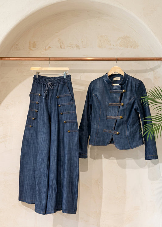 A dark blue denim two-piece outfit hanging on a wooden rack against a warm beige plaster wall. The set includes wide-leg pants with decorative button details on the legs and a matching cropped jacket with toggle closures.